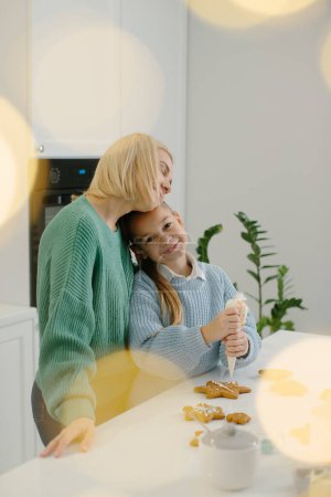 Téléchargez les photos : Une mère étreint sa petite fille alors qu'elle se tient dans la cuisine. Mère et fille décorent les biscuits de Noël en pain d'épice avec du glaçage. Concept de Noël. - en image libre de droit
