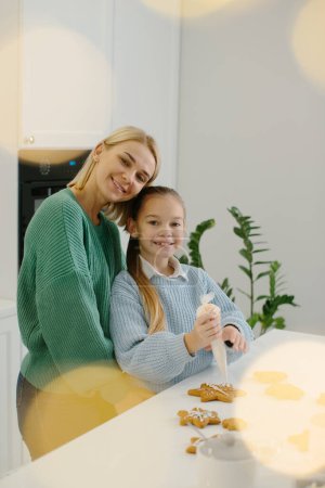 Photo for Young happy mother hugs her little daughter in the kitchen while making gingerbread cookies. - Royalty Free Image