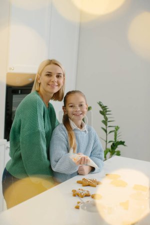 Foto de Joven madre feliz abraza a su hija pequeña en la cocina mientras hace galletas de jengibre. - Imagen libre de derechos