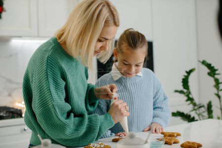 Téléchargez les photos : Mère et fille préparent des biscuits de Noël et les décorent avec du glaçage pour un joyeux Noël et des vacances heureuses - en image libre de droit
