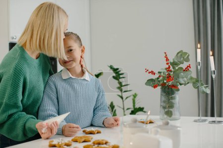 Photo for Young happy mother hugs her little daughter in the kitchen while making gingerbread cookies. - Royalty Free Image