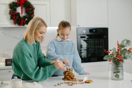 Photo for Happy mother and cute girl daughter decorating Christmas gingerbread cookies after baking while standing in cozy kitchen at home with Christmas decorations. - Royalty Free Image