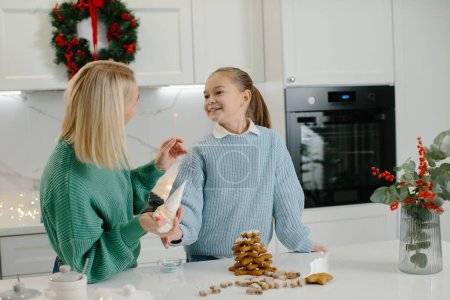 Foto de Feliz madre y linda hija niña decorando galletas de jengibre de Navidad después de hornear mientras está de pie en la acogedora cocina en casa con decoraciones de Navidad. - Imagen libre de derechos