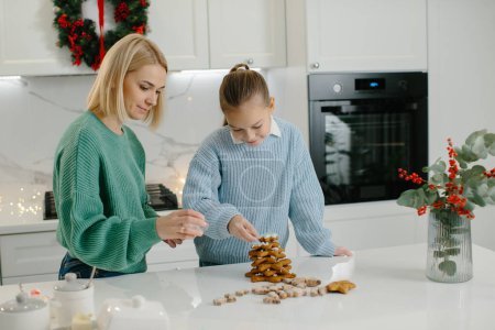 Téléchargez les photos : Mère heureuse et fille mignonne fille décorant biscuits de pain d'épice de Noël après la cuisson tout en se tenant dans la cuisine confortable à la maison avec des décorations de Noël. - en image libre de droit