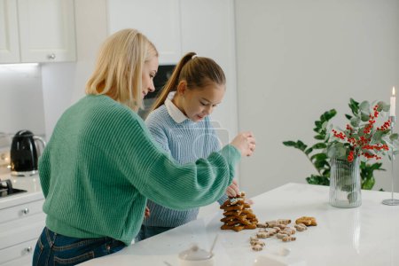Foto de Feliz madre y linda hija niña decorando galletas de jengibre de Navidad después de hornear mientras está de pie en la acogedora cocina en casa con decoraciones de Navidad. - Imagen libre de derechos