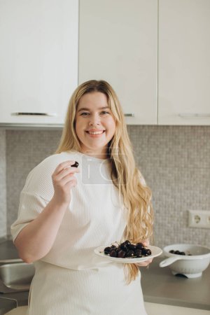 Téléchargez les photos : Jeune femme souriante mangeant des cerises fraîches dans la cuisine - en image libre de droit