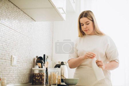 Téléchargez les photos : Jeune femme souriante dans la cuisine, faisant le petit déjeuner à partir de céréales et de lait. - en image libre de droit