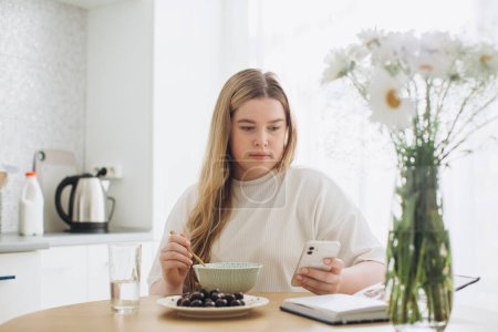 Téléchargez les photos : La fille blonde prend le petit déjeuner tout en tenant le téléphone dans ses mains. - en image libre de droit