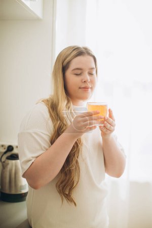 Téléchargez les photos : Portrait de jeune femme souriante tenant une tasse de thé chaud dans les mains le matin dans une cuisine - en image libre de droit