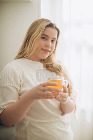 Téléchargez les photos : Portrait de jeune femme souriante tenant une tasse de thé chaud dans les mains le matin dans une cuisine - en image libre de droit