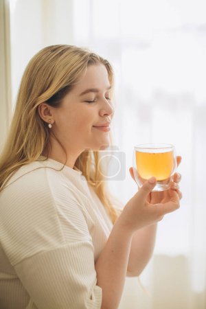 Photo for Portrait of smiling young woman holding cup of hot tea in hands in the morning in a kitchen - Royalty Free Image