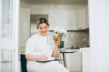 Téléchargez les photos : Jeune femme souriante buvant du thé et écrivant dans son journal intime ou journal créatif le matin dans la cuisine. - en image libre de droit
