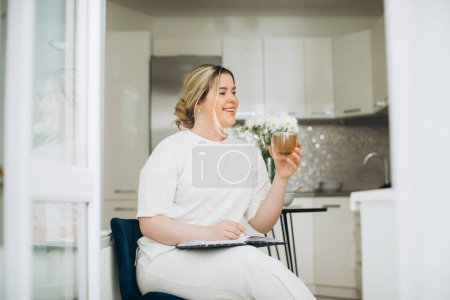 Photo for Smiling young woman drinking tea and writing in her diary or creative journal in the morning in the kitchen. - Royalty Free Image