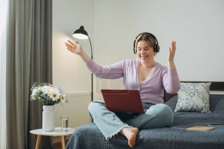 Foto de Chica con el pelo rubio usando auriculares viendo video de entretenimiento o escuchando música en el ordenador portátil mientras está sentado en la cama en el dormitorio en casa. - Imagen libre de derechos