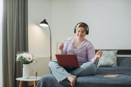 Foto de Chica con el pelo rubio usando auriculares viendo video de entretenimiento o escuchando música en el ordenador portátil mientras está sentado en la cama en el dormitorio en casa. - Imagen libre de derechos