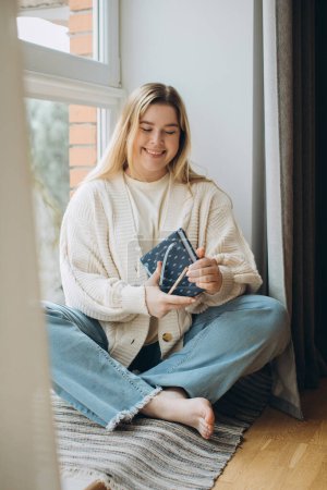Photo for A young woman is sitting at home by the window, smiling and holding a diary in her hands. - Royalty Free Image