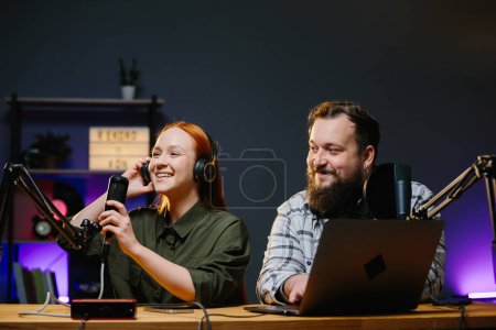 Photo for Couple of bloggers in headphones broadcasting live video, broadcasting on air, using sound studio equipment, speaking into microphone, recording video interview. - Royalty Free Image