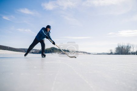 Téléchargez les photos : Un homme âgé s'entraîne à serrer la rondelle avec un bâton de hockey sur un lac gelé en hiver. - en image libre de droit