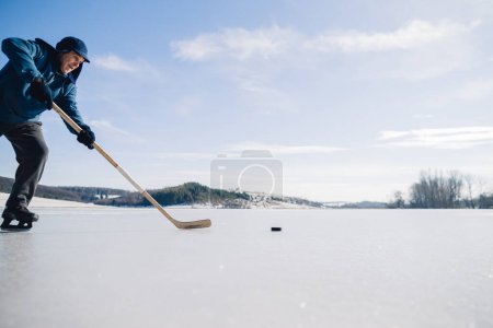 Photo for An elderly man practices stricking the puck with hockey stick on a frozen lake in winter. - Royalty Free Image