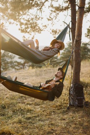 Photo for A guy and a girl are relaxing in hammocks in a pine forest at sunset. A couple in love rest in hammocks after a hike in the mountains, a guy plays the ukulele. - Royalty Free Image