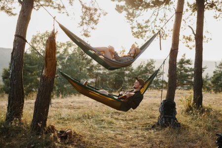 Foto de Un chico y una chica se están relajando en hamacas en un bosque de pinos al atardecer. Una pareja enamorada descansa en las hamacas después de una caminata en las montañas, un chico toca el ukelele. - Imagen libre de derechos
