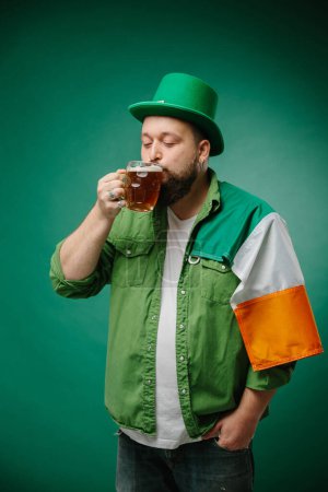 Foto de Hombre barbudo feliz con vaso de cerveza y bandera irlandesa sobre fondo verde oscuro. Día de San Patricio. - Imagen libre de derechos