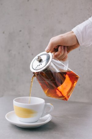 Foto de La mano de un camarero vierte té aromático en una taza, aislada sobre un fondo gris. - Imagen libre de derechos