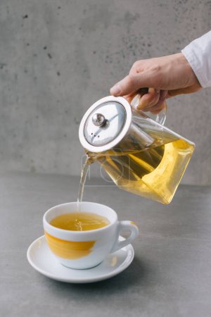 Téléchargez les photos : La main d'un barista verse du thé aromatique dans une tasse, isolé sur un fond gris. - en image libre de droit