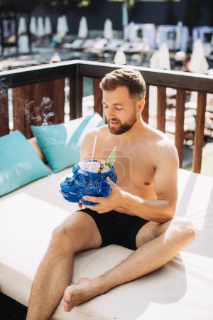 Téléchargez les photos : Beau jeune homme relaxant avec un cocktail au bord de la piscine en vacances. - en image libre de droit
