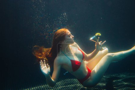 Téléchargez les photos : Belle femme rousse posant avec un cocktail sous l'eau dans une piscine. Concept vacances d'été. - en image libre de droit