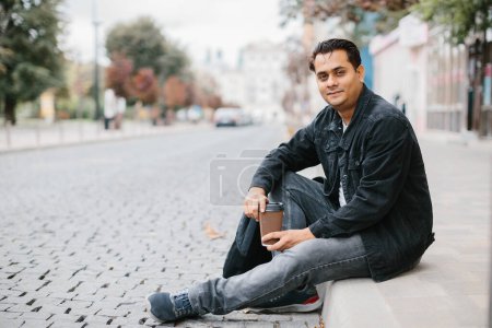 Téléchargez les photos : Un hindou souriant s'assoit par terre dans une rue de la ville et prend son café du matin. Le concept de tourisme et de mode de vie urbain. - en image libre de droit