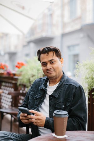 Photo for A cheerful young Indian man is drinking coffee on the terrace of a cafe and browsing social networks or chatting on his phone. - Royalty Free Image