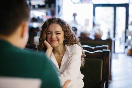 Photo for Happy young woman looking at her boyfriend with smile while both sitting in front of each other by table and enjoying date in cafe - Royalty Free Image