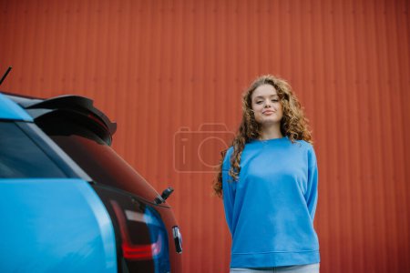Photo for A stylish young woman is standing next to her electric car in the parking lot. - Royalty Free Image