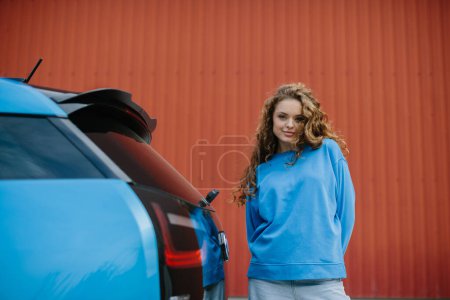 Téléchargez les photos : Une jeune femme élégante se tient à côté de sa voiture électrique dans le parking. - en image libre de droit