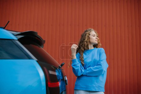 Foto de Una joven con estilo posa junto a su coche eléctrico en el estacionamiento. - Imagen libre de derechos