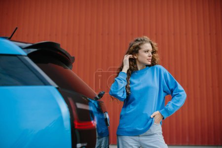 Foto de Una joven con estilo posa junto a su coche eléctrico en el estacionamiento. - Imagen libre de derechos