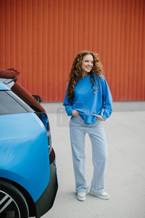 Foto de Una joven con estilo está de pie junto a su coche eléctrico en el estacionamiento. - Imagen libre de derechos