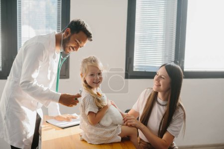 Téléchargez les photos : Une petite fille d'âge préscolaire avec sa mère est examinée par un pédiatre. Un jeune homme, un pneumologue, effectue un examen à l'aide d'un phalendoscope. - en image libre de droit