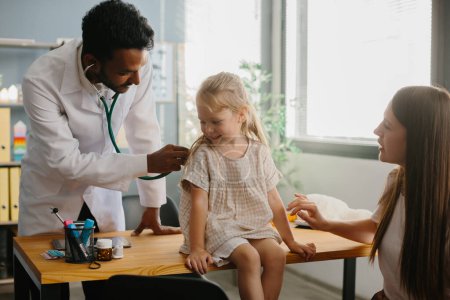 Photo for Mother and child seeing family practitioner. Smiling pediatrician with stethoscope checking little girl's lungs. - Royalty Free Image