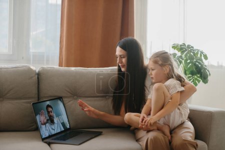 Téléchargez les photos : Une jeune mère avec sa petite fille consulte son médecin de famille en ligne à l'aide d'un ordinateur portable. - en image libre de droit