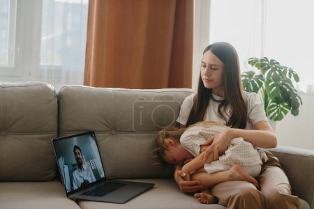 Photo for A young mother with her little daughter consults with her family doctor online using a laptop. - Royalty Free Image