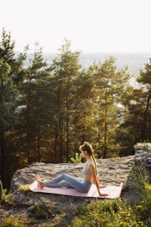 Téléchargez les photos : Fit jeune femme pratique le yoga sur les rochers sur le fond de la forêt de pins. Une belle fille médite et fait des exercices de respiration dans les montagnes. - en image libre de droit