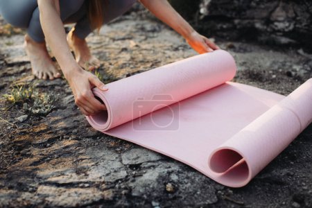 Téléchargez les photos : Une jeune femme en vêtements de fitness roule son tapis de yoga après une séance d'entraînement dans un bel endroit de montagne. - en image libre de droit