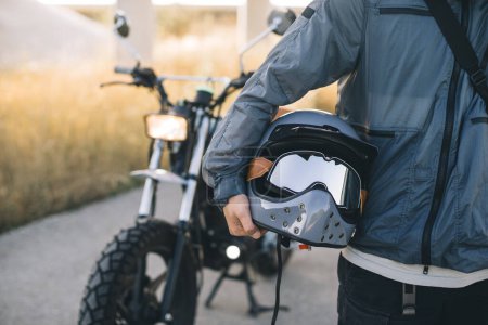Téléchargez les photos : Un motard tient un casque dans ses mains sur le fond d'une moto au coucher du soleil. - en image libre de droit