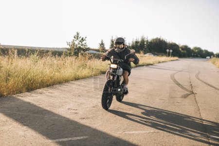 Téléchargez les photos : Un homme avec une moto vintage sur l'autoroute au coucher du soleil. Un homme conduit un coureur de café. Concept de style de vie motard - en image libre de droit