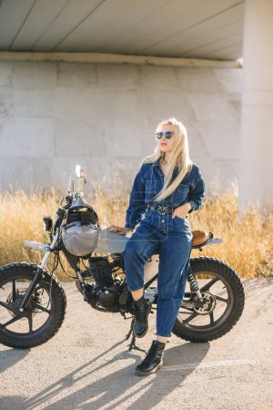 Photo for A young blonde woman in denim clothes and sunglasses poses on a motorcycle at sunset. - Royalty Free Image