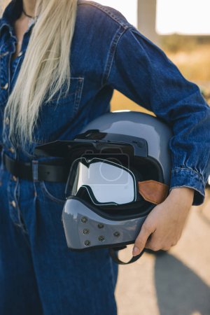 Téléchargez les photos : Une jeune femme blonde tient un casque de protection dans ses mains sur le fond d'une moto au coucher du soleil. - en image libre de droit