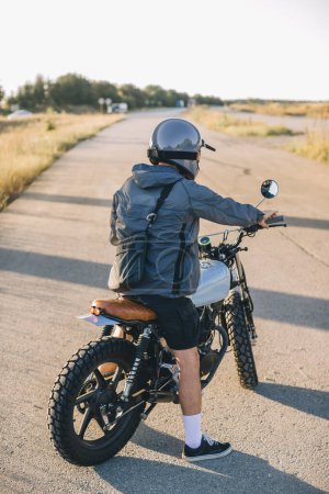 Téléchargez les photos : Biker sur une moto de course de café sur la route au coucher du soleil. Vue arrière. - en image libre de droit