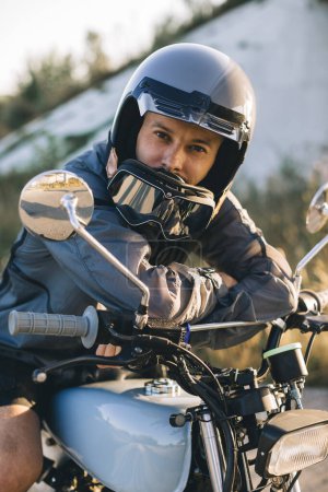 Téléchargez les photos : Un jeune motard élégant est assis sur une moto sur la route contre le coucher du soleil. Un regard sur la caméra. - en image libre de droit